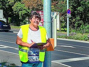 Frau mit gelber Warnweste und Schreibzeug steht neben Akustikampel