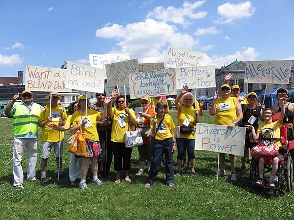 Eine große Gruppe von 16 Personen in gelben T-Shirts halten Schilder mit diversen Sprüchen für Inklusion in die Luft. 