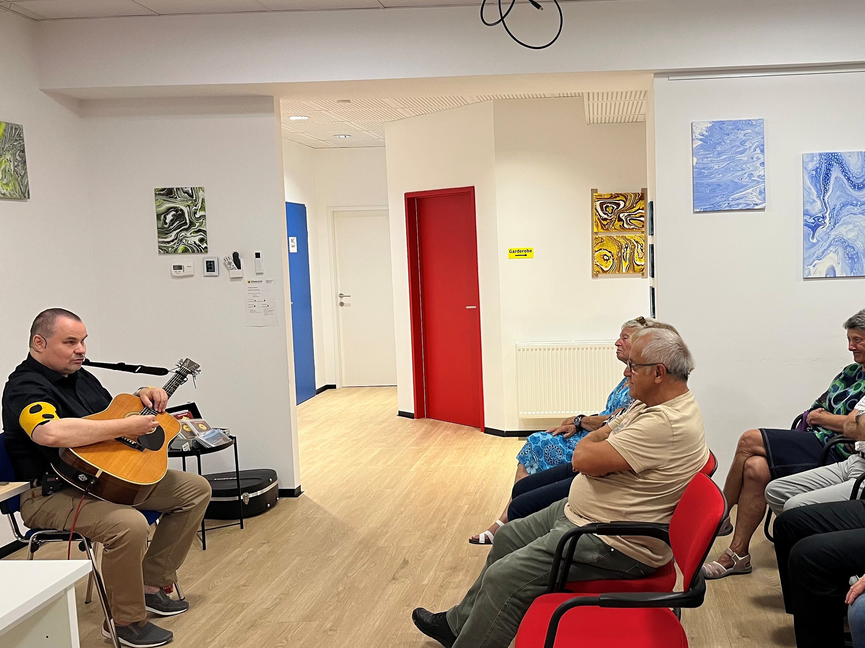 Foto: © Hlava Mann spielt in einem hellen Raum Gitarre, während mehrere Personen auf Stühlen sitzen und ihm zuhören. An den Wänden hängen abstrakte Bilder; im Hintergrund sind rote Türen zu sehen.