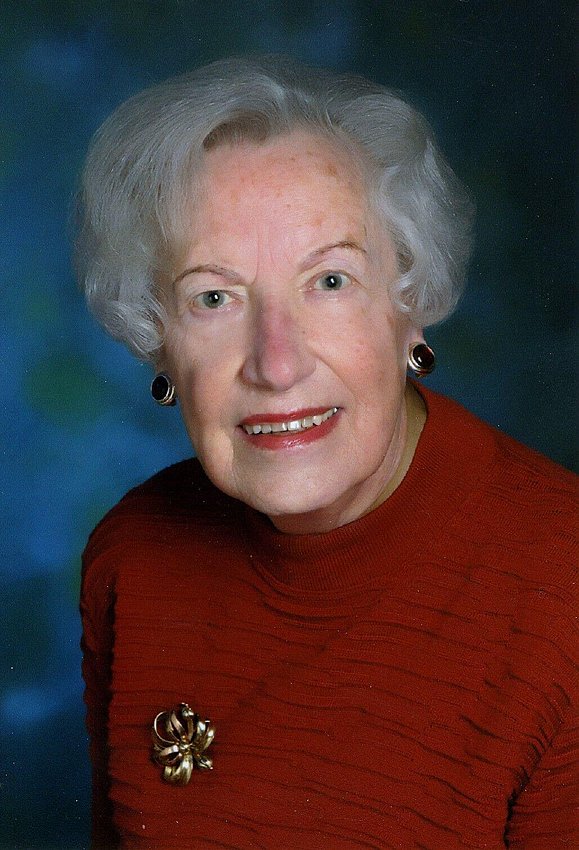 Eine ältere Dame mit grauer Dauerwelle, großen Ohrringen und rotem Lippenstift lächelt in die Kamera.