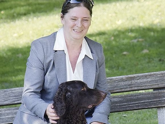 Blindenführhund Daphne, ein Curly Coated Retriever mit Blindenführhundgeschirr, sitzt vor einer Bank auf der ihr blindes Frauchen sitzt.