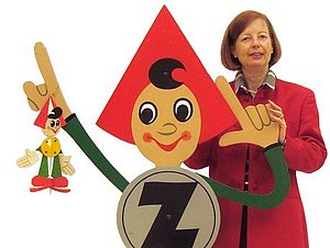 Die Werbefigur "Sparefroh" in Lebensgröße zeigt mit beiden Händen nach links oben, an einer Hand hängt ein kleinerer Sparefroh. Hinter ihm steht eine Frau in rotem Blazer.