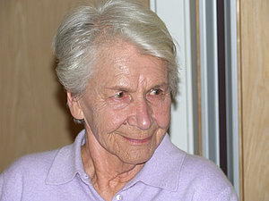 Eine ältere Dame mit kurzen weißen Haaren blickt leicht lächelnd an der Kamera vorbei