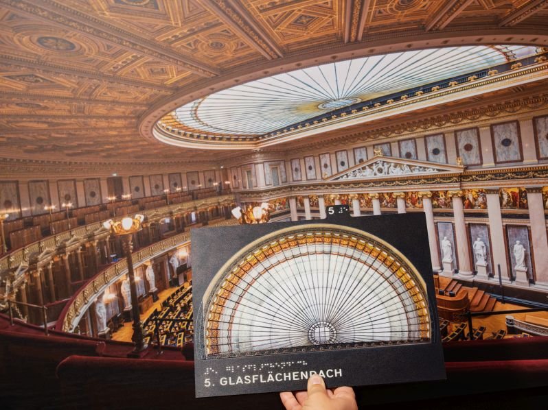 Man sieht eine tastbare Tafel, im Hintergrund einen Saal im Parlament