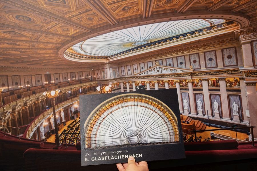 Man sieht eine tastbare Tafel, im Hintergrund einen Saal im Parlament