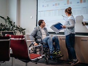 Mann mit Rollstuhl und stehende Frau im Büro schauen sich Zahlen auf einer Leinwand an