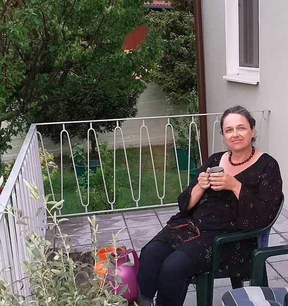 Unser Mitglied Céleste sitzt mit einer Kaffeetasse in der Hand auf einem Balkon. Sie trägt dunkle Kleidung und hat dunkles Haar. Auf dem Balkon steht ein 2. Stuhl, einige Pflanzen und Gießkannen. 