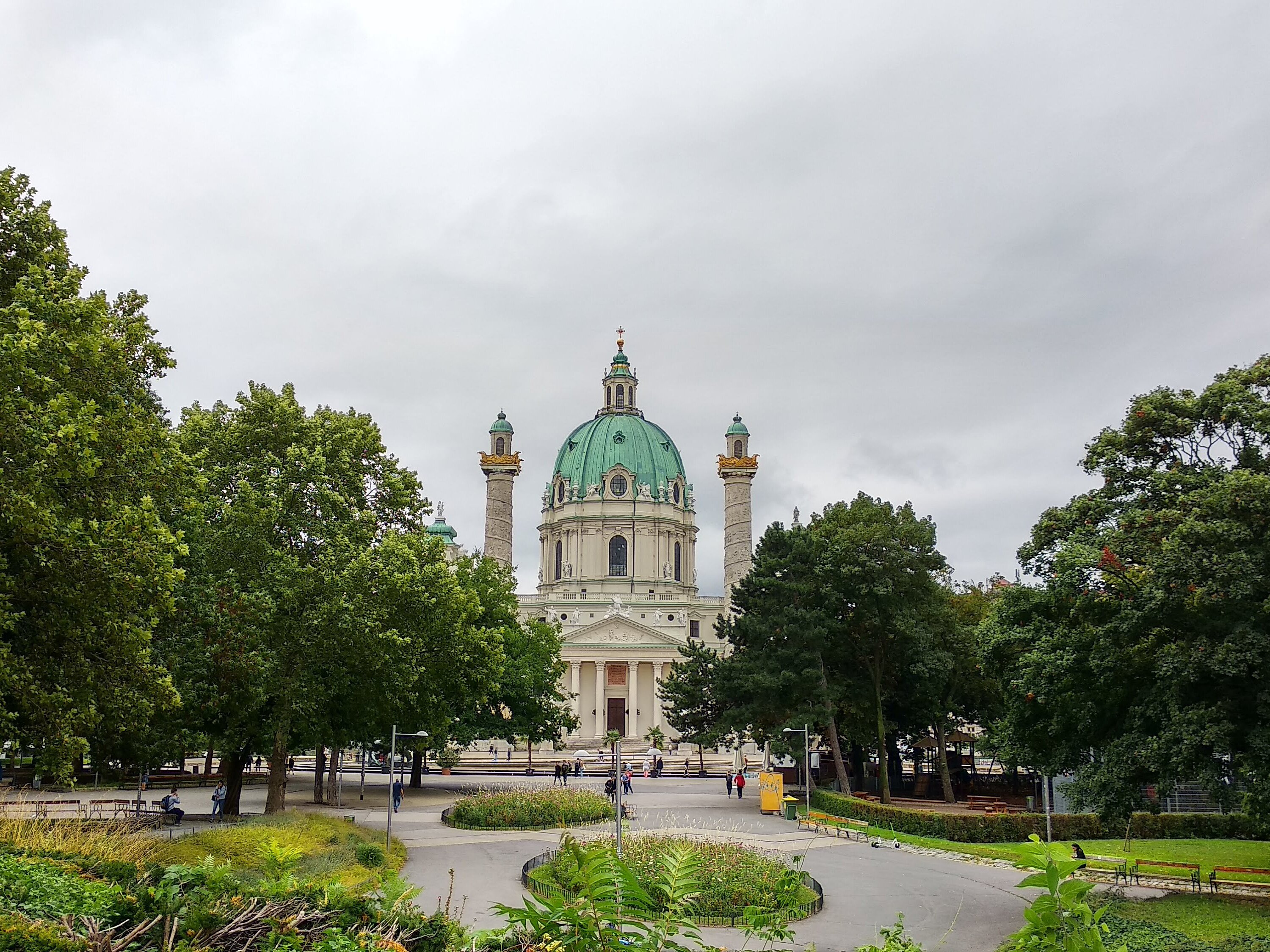 Blick durch einen Park auf eine große Kirche mit grüner Kuppel und zwei Türmen unter grauem Himmel.