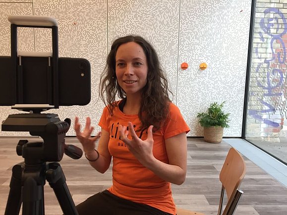 Trainerin Nadine erzählt bei einer Live-Aufzeichnung über das Wesen des karibischen Tanzes Zouk. Copyright: Lillis Ballroom Eine junge Frau mit Locken sitzt vor einem Telefon auf einem Stativ und erklärt gestikulierend in die Kamera
