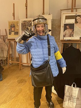 Kunsthistorisches Museum ist mit seinen Tastführungen bei unseren Mitgliedern sehr beliebt! ältere Frau mit Ritterhelm und blauer Jacke mit gelber Blindenkennzeichnung steht vor Bildern