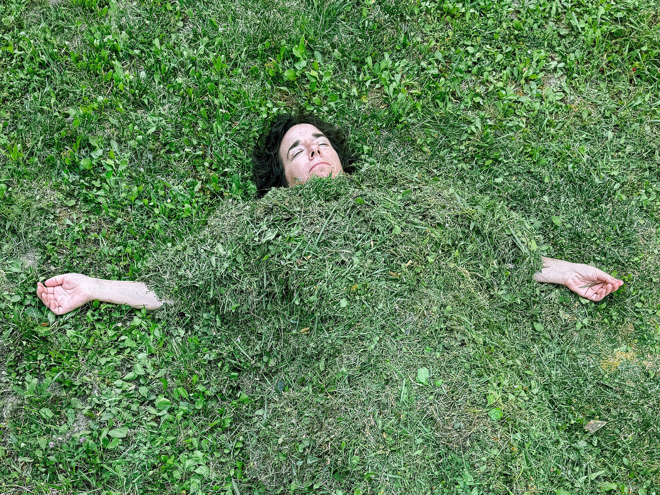 Person liegt auf einer Wiese und ist bis zum Hals mit Gras bedeckt; nur das Gesicht und die ausgestreckten Arme sind sichtbar.