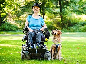 Junge Frau im Rollstuhl auf einer grünen Wiese, neben ihr ein Assistenzhund.