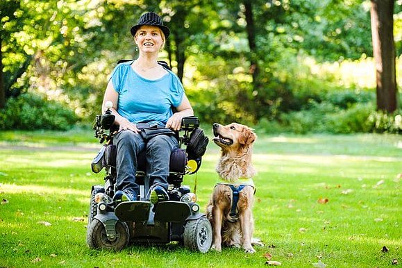 Junge Frau im Rollstuhl auf einer grünen Wiese, neben ihr ein Assistenzhund.