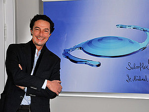 Ein Mann steht lächelnd vor einem Plakat auf dem eine neue Sulcoflex-Linse vorgestellt wird.