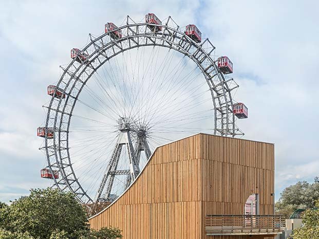 Das Riesenrad ist über Baumwipfeln und hinter einer Holzkonstruktion vor bewölktem Himmel zu sehen.