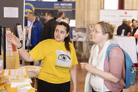 Junge Frau mit gelbem Shirt mit Auge drauf zeigt anderer Frau mit Rucksack bei einem Messestand etwas am Messestand