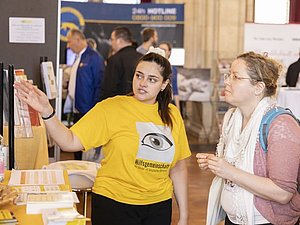 Junge Frau mit gelbem Shirt mit Auge drauf zeigt anderer Frau mit Rucksack bei einem Messestand etwas am Messestand