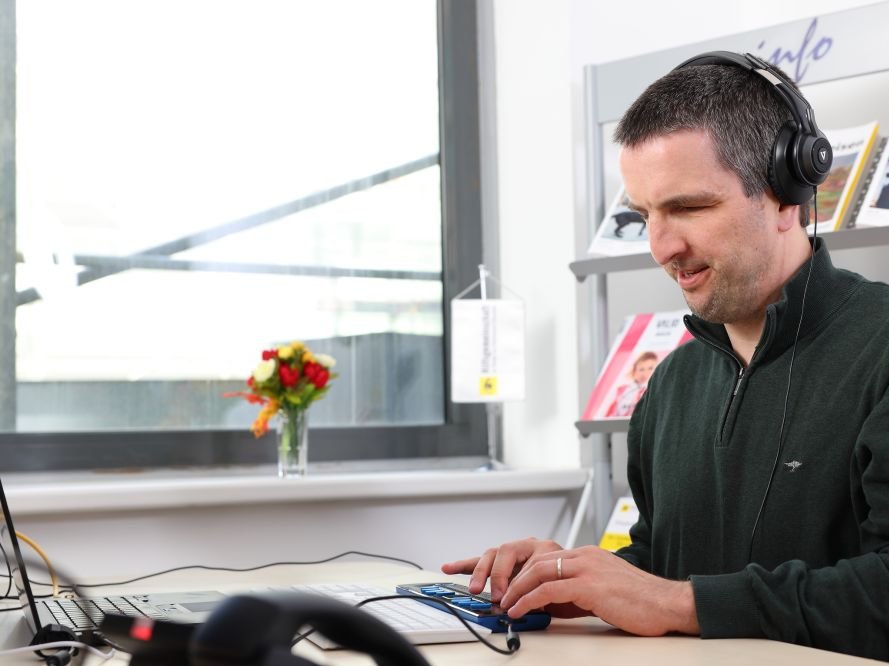 Mann mit schwarzem Pullover sitzt mit Kopfhörern am Laptop und tastet auf Braillezeile