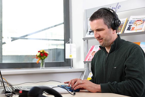 Mann mit schwarzem Pullover sitzt mit Kopfhörern am Laptop und tastet auf Braillezeile