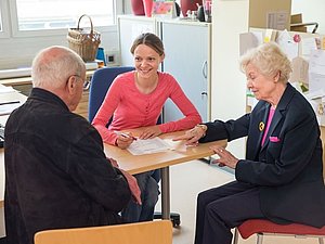 Ein älteres Paar sitzen nebeneinander, einer jungen frau zugewandt und betrachten ein Blatt Papier das zwischen ihnen auf einem Tisch liegt, die ältere Frau trägt einen Blinde-Button