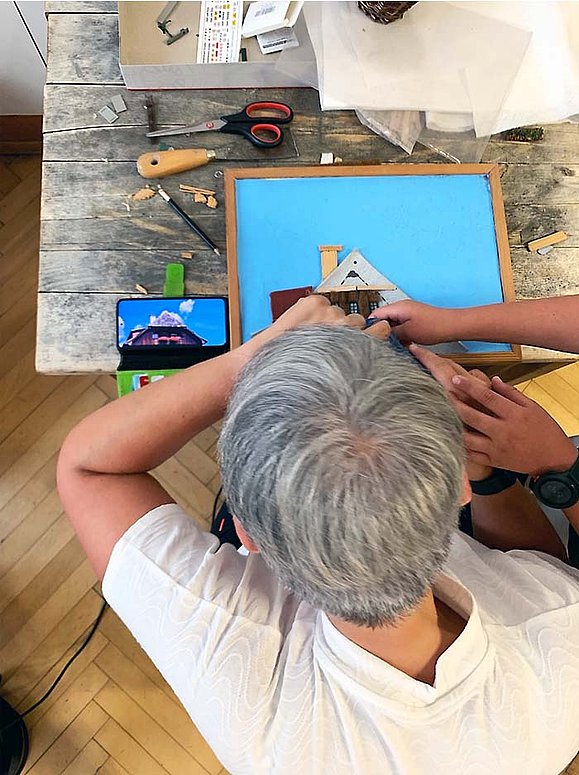 Eine Person mit kurzen grauen Haaren ist über einen Werktisch gebeugt auf dem diverse Werkzeuge und Materialien liegen. In einem Rahmen setzt die Person eine taktile Fotografie zusammen, die Vorlage wird auf einem daneben liegenden Handy angezeigt. 