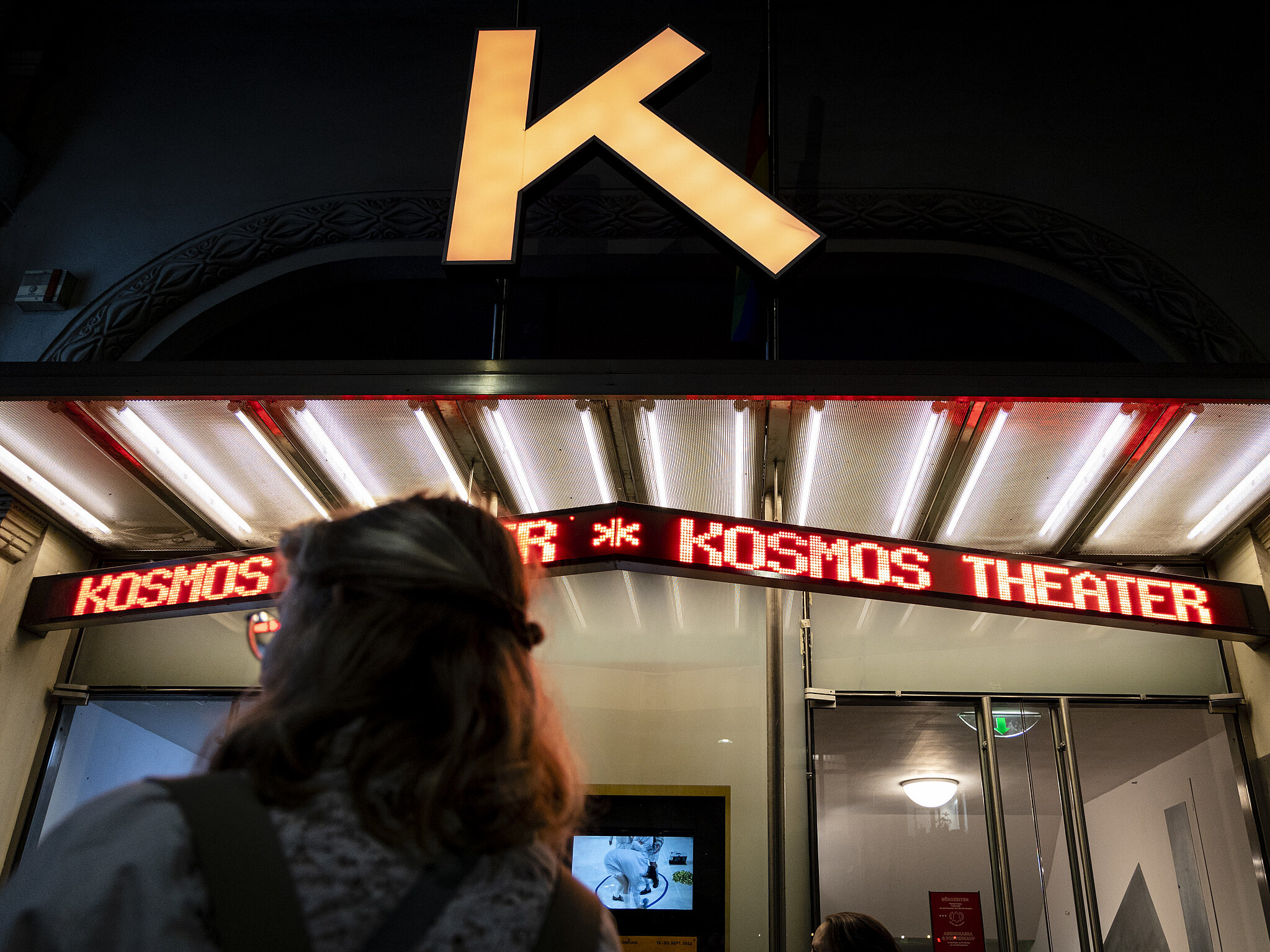 Nächtliche Aufnahme des Eingangs zum Kosmos Theater mit leuchtendem rotem Schriftzug; im Vordergrund steht eine Person mit dem Rücken zur Kamera. Above dem Eingang ist ein großes beleuchtetes „K“-Symbol zu sehen.
