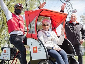 Fahrrand mit Sitzmöglichkeit vorne vor Prater: Junge Person mit roter Weste sitzt auf Fahrrad, vorn drin sitzt winkende ältere Dame, dahinter posiert älterer Herr