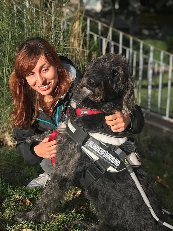 Frau mit langen braunen Haaren lächelt in die Kamera und umarmt schwarzen Hund mit krausen Haaren. Der Hund trägt ein Geschirr mit der Aufschrift: Blindenführhund