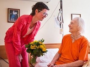 Eine Testamentsspenderin sitzt auf einem Bett. Von links beugt sich eine Mitarbeiterin mit einem Blumenstrauß zu ihr hin.