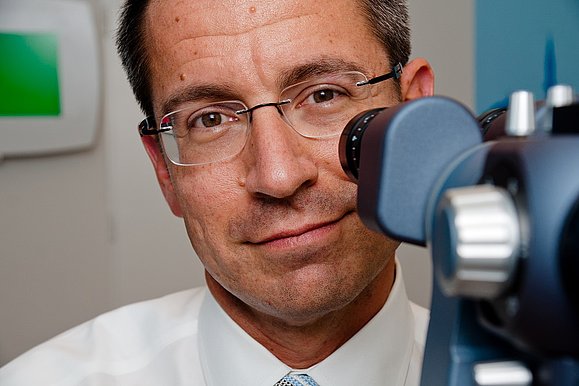 Bild eines Arztes der in die Kamera lächelt, vor ihm steht ein Gerät zur Augenuntersuchung