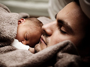 Ein Mann liegt mit einem Baby auf der Brust in eine braune Decke gekuschelt.