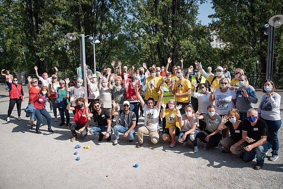 Mehrere Menschen stehen im Sommer auf einem sandigen Platz, hinter ihnen sind einige Bäume zu sehen, manche winken in die Kamera.