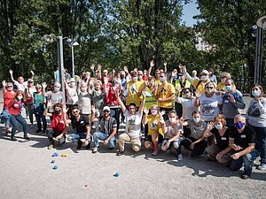 Mehrere Menschen stehen im Sommer auf einem sandigen Platz, hinter ihnen sind einige Bäume zu sehen, manche winken in die Kamera.