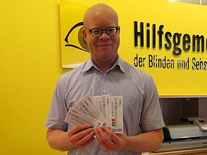 Ein Mann mit Brille und Glatze hält 5 REWE Einkaufsgutscheine in den Händen. Hinter ihm sieht man das Hilfsgemeinschaft-Logo.
