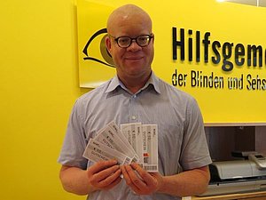 Ein Mann mit Brille und Glatze hält 5 REWE Einkaufsgutscheine in den Händen. Hinter ihm sieht man das Hilfsgemeinschaft-Logo.