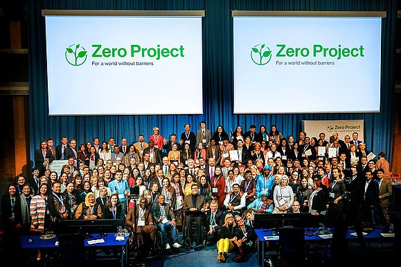 Viele Menschen stehen auf einer Bühne nebeneinander. Darüber sieht man 2x das Zero Project Logo auf einer Leinwand.