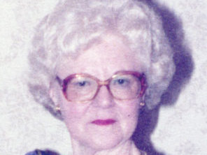 Eine ältere Dame mit Dauerwelle, großer Brille und rotem Lippenstift blickt leicht lächelnd in die Kamera.