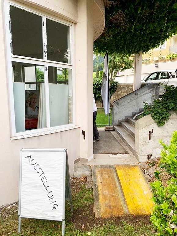 Foto eines Gebäudeeingangs. Von der Straße führen Stufen hinunter, seitlich, dem Betrachter zugewandt, ist eine Rasenfläche, die an den Eingang grenzt. Hier geht eine Stufe hinauf, die mit einer Rampe aus gelbem Holz überbrückt wurde. Daneben steht ein Schild, auf dem handgeschrieben "Ausstellung" steht. 