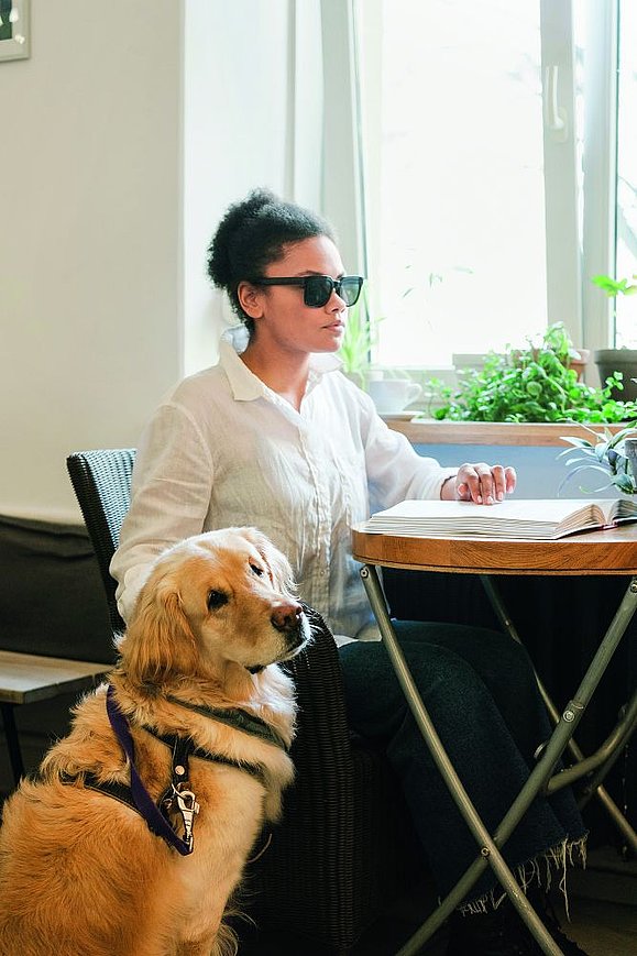 Blinde Frau sitzt an Tisch und liest Braille mit einer Hand und andere Hand auf Hund