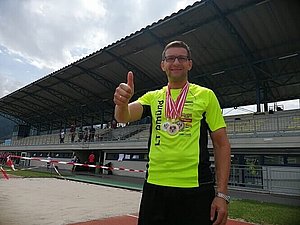 Ein Mann steht mit neongelben T-Shirt und drei Medaillen um den Hals in einem Stadion. Er hält den erhobenen Daumen in die Kamera. 