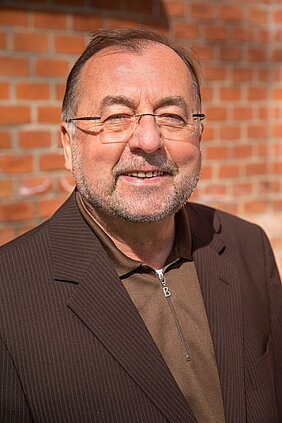 älterer Mann mit brauner Jacke und Brille vor roter Backsteinwand lächelt in die Kamera