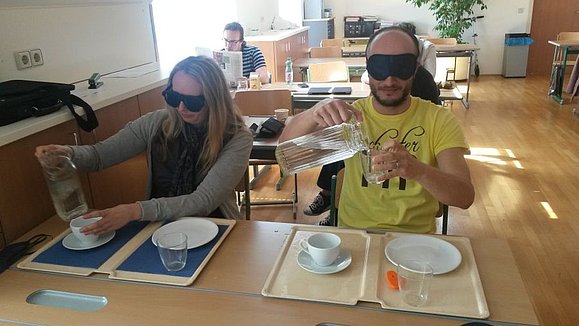 Eine Frau und ein Mann mit Simulationsbrillen sitzen nebeneinander an einem Tisch. Beide haben ein Essenstablet mit unserschiedlichen Kontrasten vor sich stehen und versuchen Wasser in ein Glas einzuschenken.