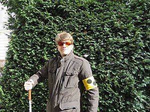 Mitglied Max steht mit Blindenschleife, Kantenfilterbrille und Langstock vor einem Busch.