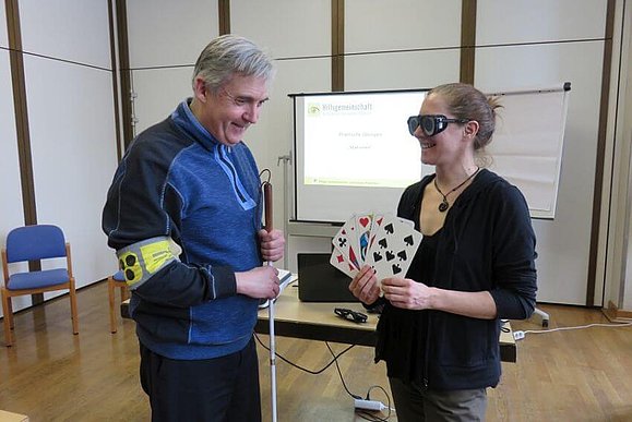 Petra Wrba hält große Tarockkarten in den Händen, sie trägt dabei eine Simulationsbrille. Harald Rother steht mit Taststock und Armbinde daneben.