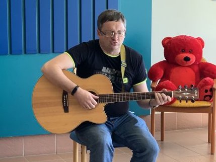 Ruslan, ein hochgradig sehbehinderter Musikliebhaber aus der Ukraine, der derzeit in der Waldpension lebt, sitzt auf einem Schemel und spielt Gitarre vor einer blauen Wand. Neben ihm sitzt ein roter Teddybär auf einem Sessel.