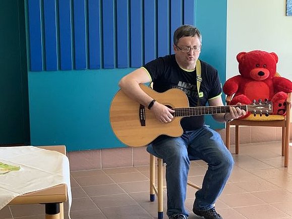 Ruslan, ein hochgradig sehbehinderter Musikliebhaber aus der Ukraine, der derzeit in der Waldpension lebt, sitzt auf einem Schemel und spielt Gitarre vor einer blauen Wand. Neben ihm sitzt ein roter Teddybär auf einem Sessel.