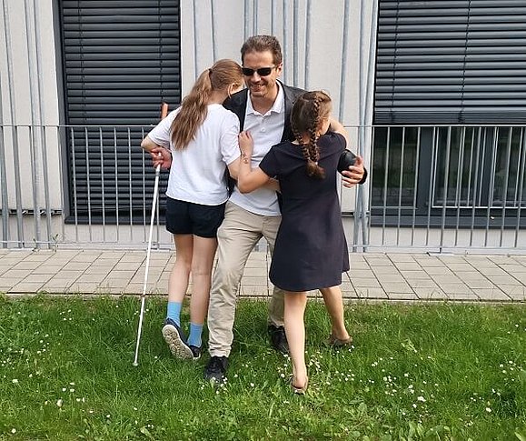 Michele wird von seinen Töchtern umarmt. Er steht mit Langstock und Brille in einer Wiese, links und rechts in seinen Armen seine Töchter.