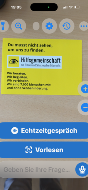 Smartphone-App zeigt ein gelbes Infoplakat der Hilfsgemeinschaft für Blinde und Sehschwache sowie Schaltflächen zum Vorlesen und für Echtzeitgespräche.