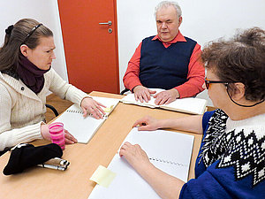 Kursleiter und zwei Schülerinnen sitzen an einem Tisch und betasten Seiten in Blindenschriftbüchern