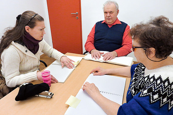 Kursleiter und zwei Schülerinnen sitzen an einem Tisch und betasten Seiten in Blindenschriftbüchern